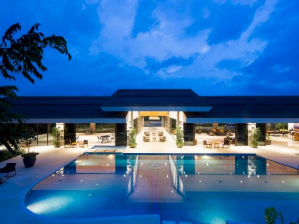 A luxurious, well-lit building with a large swimming pool in front, surrounded by outdoor seating under a twilight sky.