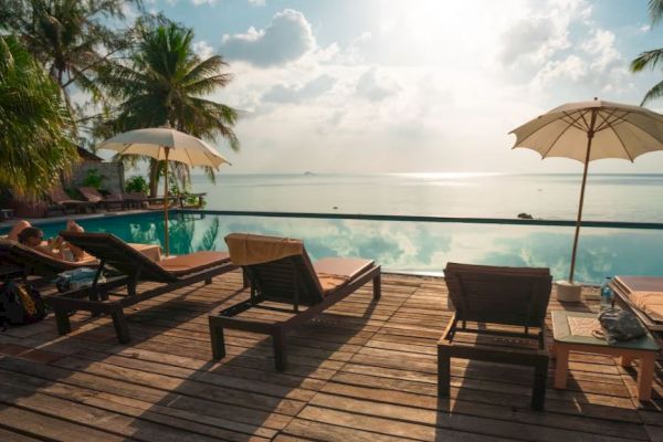 Poolside scene with loungers and umbrellas, overlooking a serene body of water under a sunny sky, surrounded by palm trees.