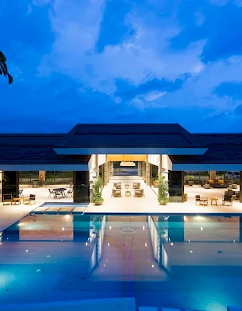 The image shows a luxurious building with a large, illuminated swimming pool under a blue twilight sky.