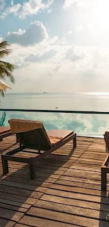 A serene poolside scene with lounge chairs, umbrellas, palm trees, and a view of the ocean under a partly cloudy sky.