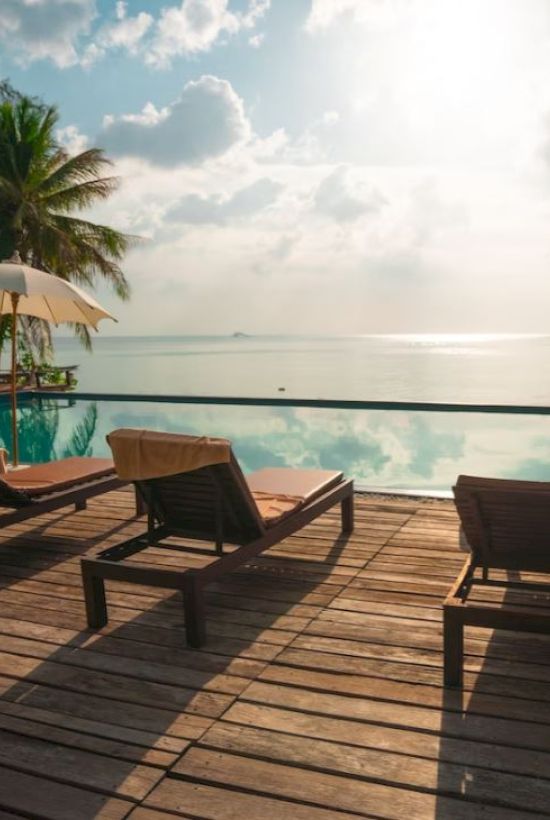 Ocean view with lounge chairs and umbrellas on a wooden deck by the water, surrounded by palm trees under a partly cloudy sky.