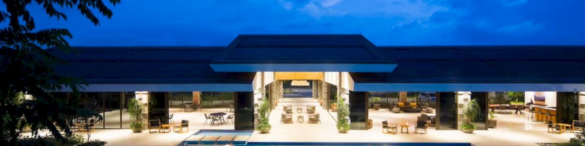 The image shows a modern building with large, open spaces and a swimming pool in front, set against an evening sky.