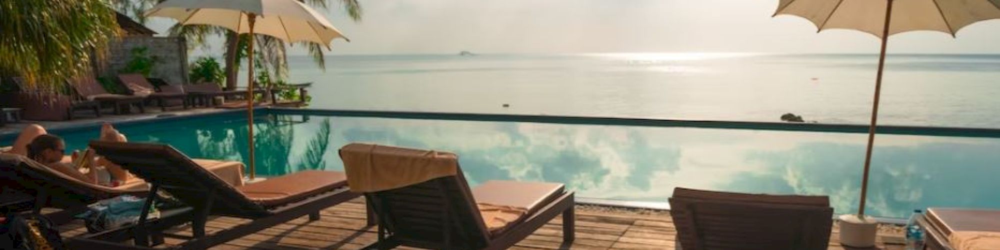 Beachside pool with lounge chairs, umbrellas, and palm trees. Overlooks a calm ocean under a partly cloudy sky, creating a tranquil scene.