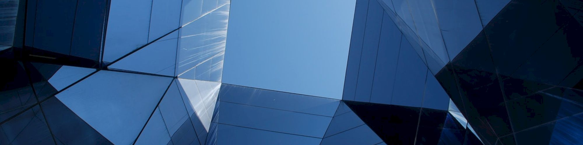The image shows a geometric view looking up at tall, reflective glass buildings with a clear blue sky at the center.