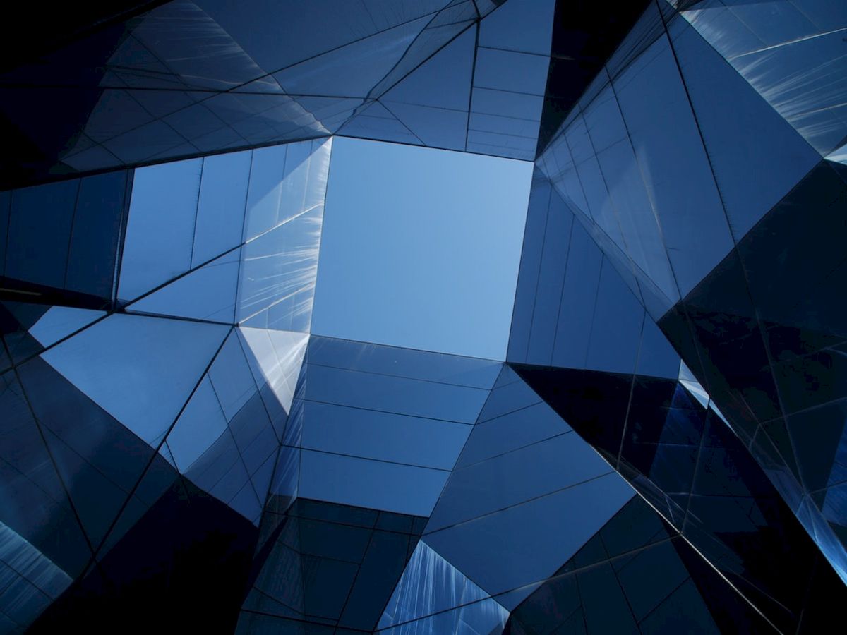 A geometric view looking up at reflective glass architecture against a clear blue sky, creating abstract patterns and angles.