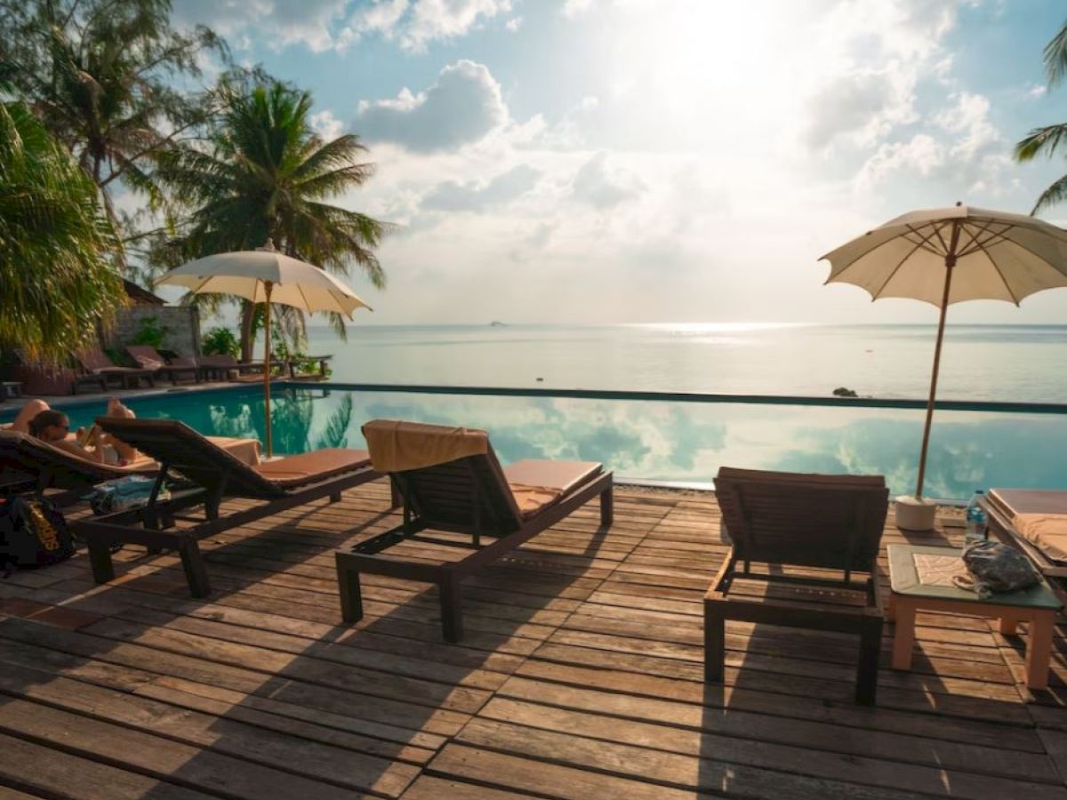 Poolside scene with lounge chairs, umbrellas, and a palm tree, overlooking a calm sea under a bright sky.