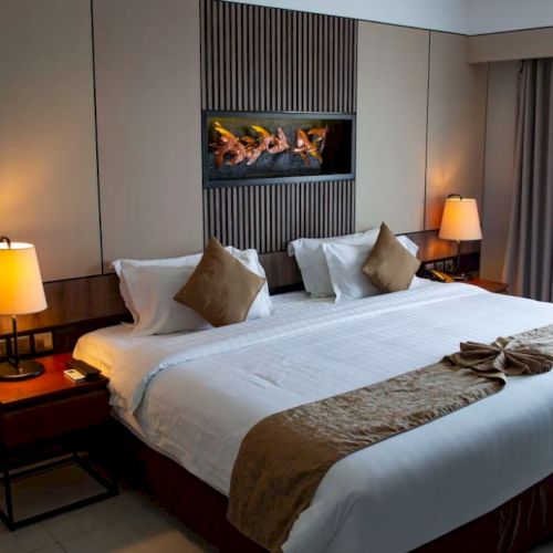 A modern hotel room with a large bed, decorative pillows, two side tables with lamps, and a wall-mounted artwork above the headboard.