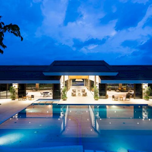 A luxurious poolside area with a modern building in the background, beautifully lit under a blue evening sky.