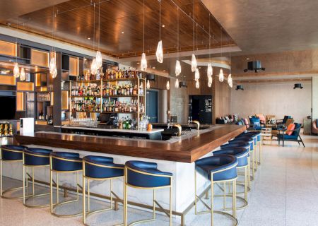 A modern bar with blue stools, wooden surfaces, and hanging lights. The shelves are stocked with various bottles. Cozy seating is in the background.