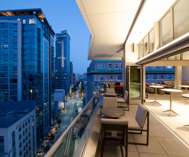 A modern rooftop terrace overlooking a city street, lined with tall buildings and featuring outdoor seating with tables.