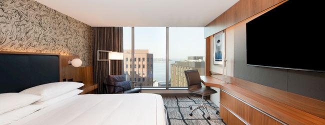 The image shows a modern hotel room with a large bed, a flat-screen TV, a desk, a chair, and a window offering a view of city buildings.