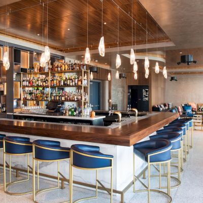 A modern bar with blue-cushioned stools, a well-stocked back bar, pendant lights, and a cozy seating area in a stylish, open space.