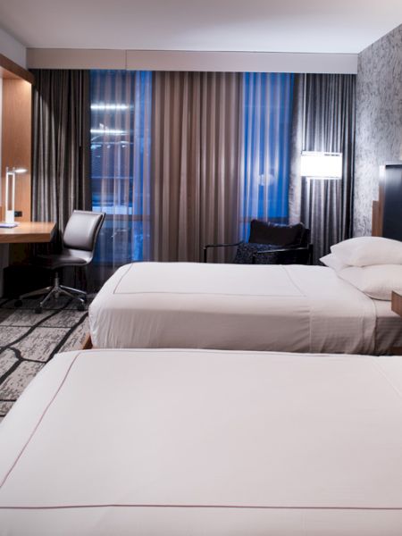 A modern hotel room with two beds, a TV, desk, and lamp. The room has stylish decor with patterned wallpaper and large windows.