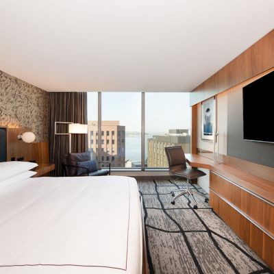 A modern hotel room with a large bed, desk, chair, TV, and window view of buildings. Clean, minimalist design with wooden accents.