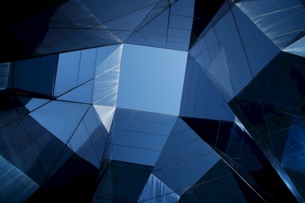 A geometric view upwards through a structure of mirrored glass panels, reflecting blue sky and clouds, creating an abstract, modern effect.