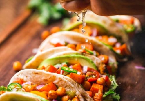 Tacos with chickpeas, avocado, and vegetables on a wooden surface, with a hand squeezing lime over them.