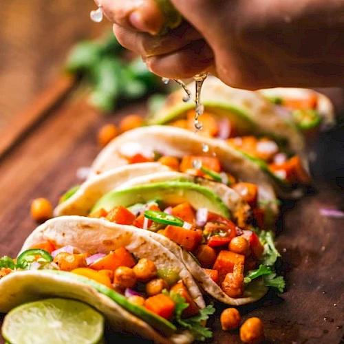 Tacos with chickpeas, avocado, and vegetables on a wooden surface, with a hand squeezing lime over them.