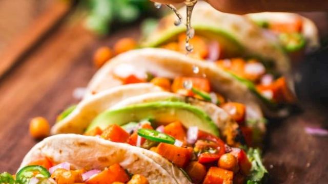 Tacos with chickpeas, avocado, and vegetables on a wooden surface, with a hand squeezing lime over them.