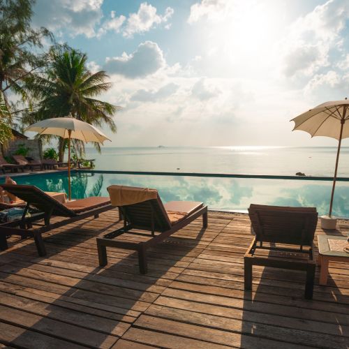 The image shows a tropical poolside setting with lounge chairs and umbrellas, overlooking a calm sea under a cloudy sky and bright sun.