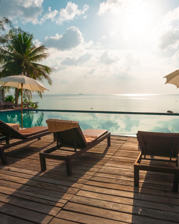 The image shows a tropical poolside setting with lounge chairs and umbrellas, overlooking a calm sea under a cloudy sky and bright sun.