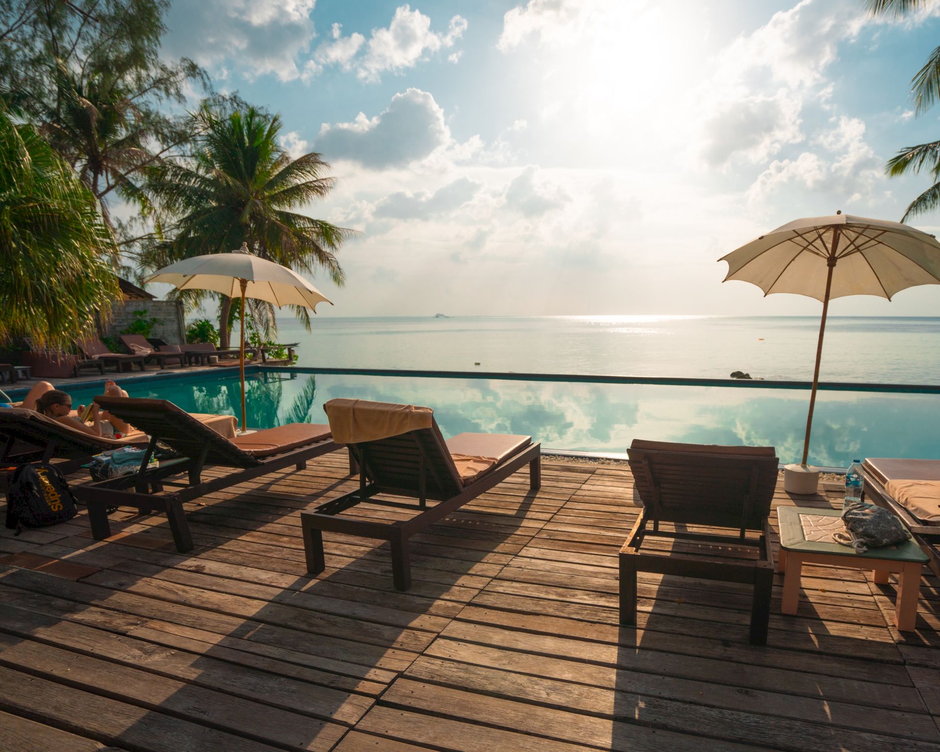 A serene poolside scene with lounge chairs, umbrellas, palm trees, and a sunny view of the ocean.