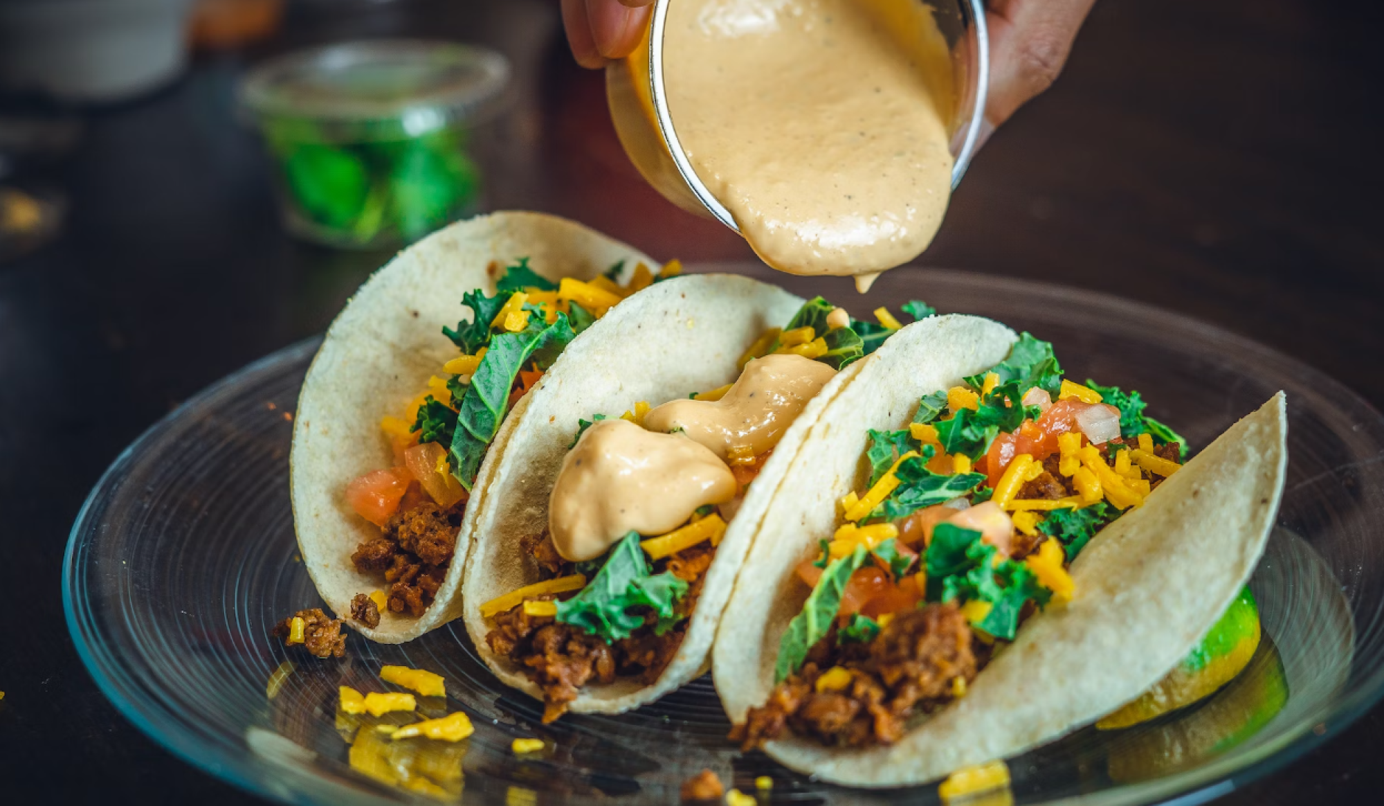 Three tacos with toppings like cheese, tomato, and greens, drizzled with a creamy sauce on a clear plate.