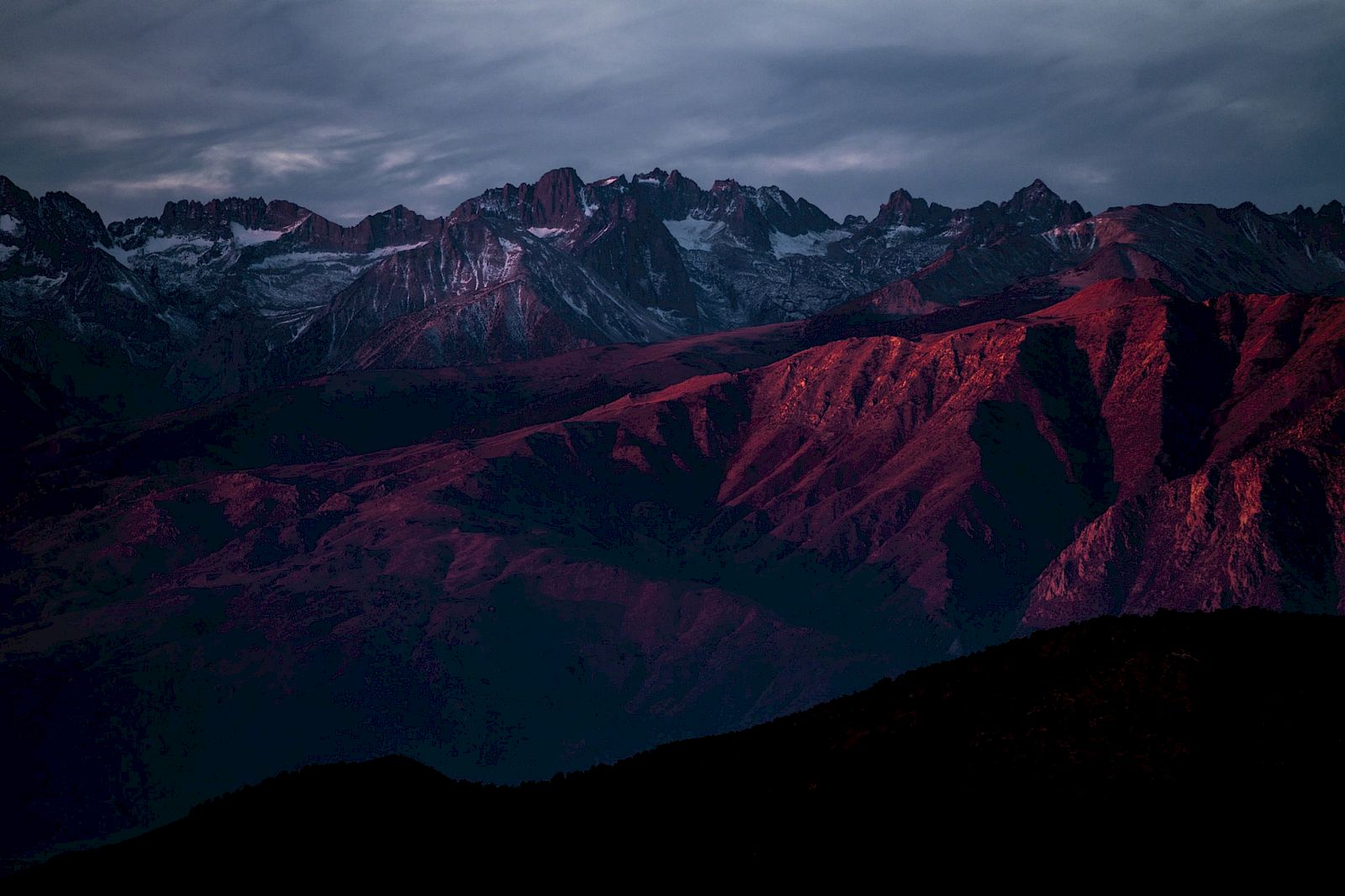 A mountain range bathed in red and purple light under a cloudy sky.