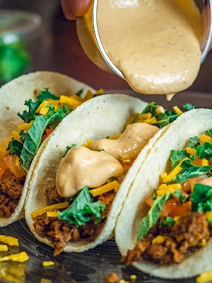 Three tacos with ground meat, lettuce, cheese, and sauce being poured on top, served on a plate.