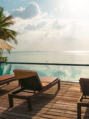 The image shows a serene poolside scene with loungers, umbrellas, and an infinity pool overlooking a calm ocean under a partly cloudy sky.