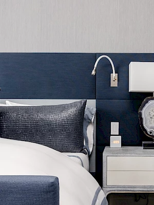 A modern bedroom setup with a large bed, blue headboard, decorative pillows, and a nightstand holding a lamp and books.