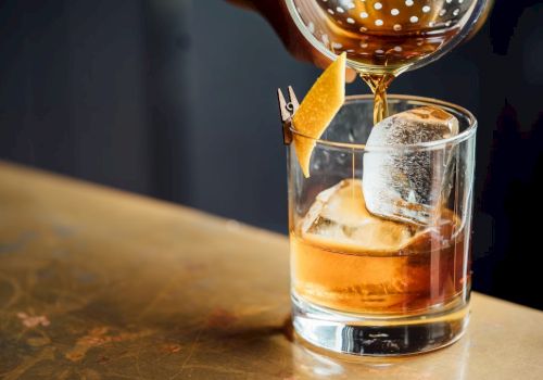A cocktail is being poured into a glass with ice and a lemon peel garnish rests on the side, set on a wooden surface.