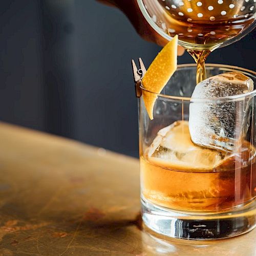 A cocktail is being poured into a glass with ice and a lemon peel garnish rests on the side, set on a wooden surface.