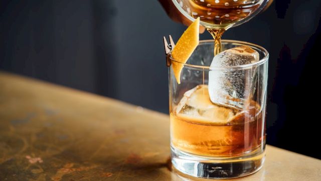 A cocktail is being poured into a glass with ice and a lemon peel garnish rests on the side, set on a wooden surface.