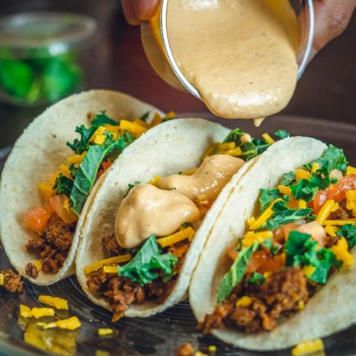 Three tacos with ground meat, cheese, and greens being topped with a creamy sauce on a plate.