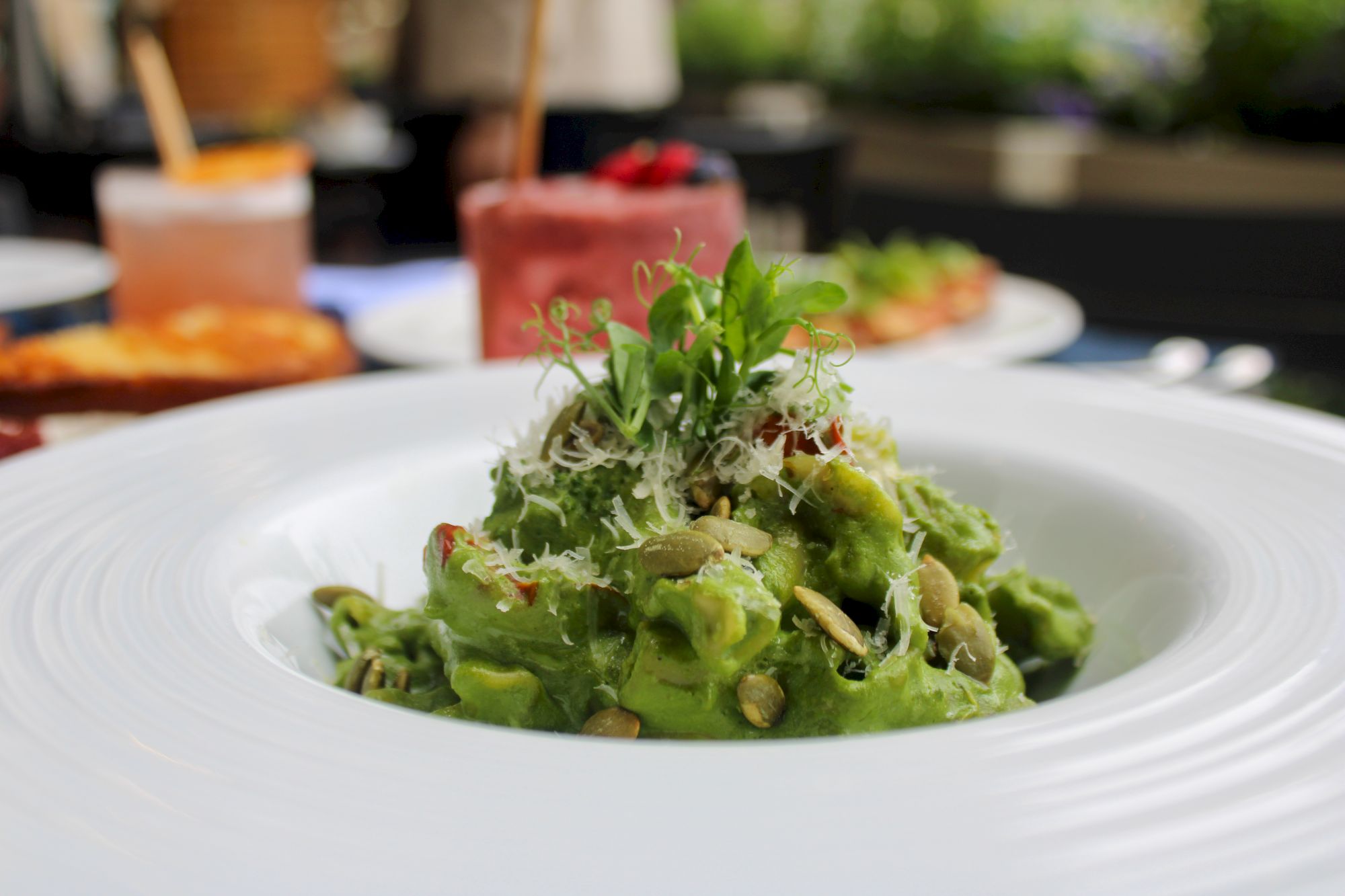 A plate of pasta with green sauce, garnished with seeds and herbs, is in focus with other dishes blurred in the background.