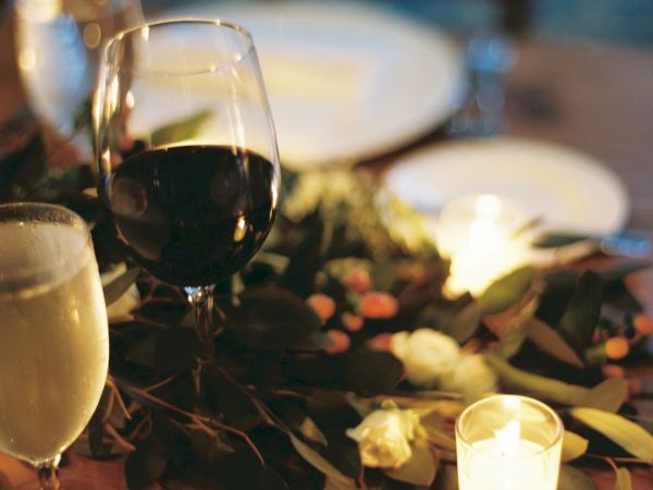 A table with a wine glass, beer, greenery, a lit candle, and plates, set for a cozy dinner.