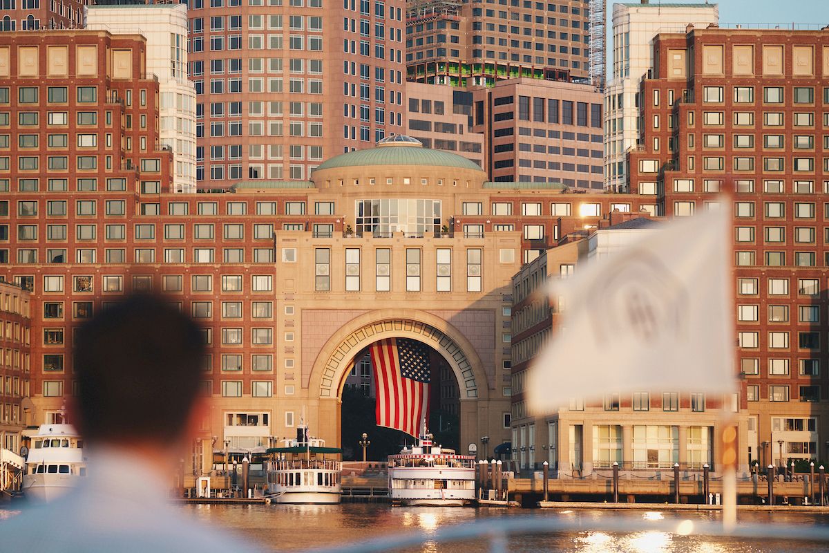 The image shows a patriotic scene with a large American flag hanging in a building archway, surrounded by cityscape and waterfront views.
