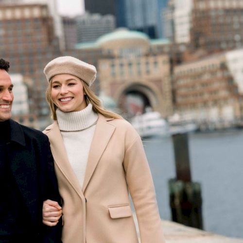 A couple walking by a waterfront with a cityscape in the background, both smiling and dressed warmly.
