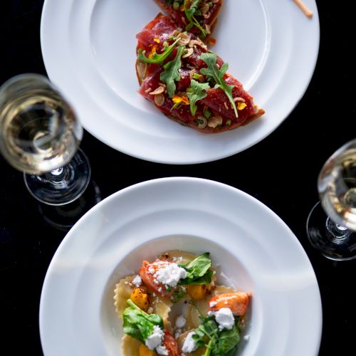 Two plated dishes overhead: a taco-like item with cilantro, chile glaze, and sticks on a white plate, and a creamy bowl with vegetables and herbs.