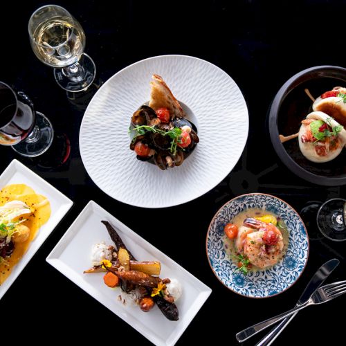 A stylish spread of assorted plated dishes and drinks on a black background, featuring small bites, sauces, and wine glasses.