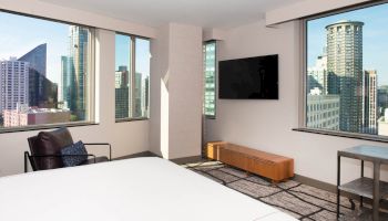 A modern hotel room with large windows shows a city skyline. There's a bed, TV, chair, and a small table in the room.