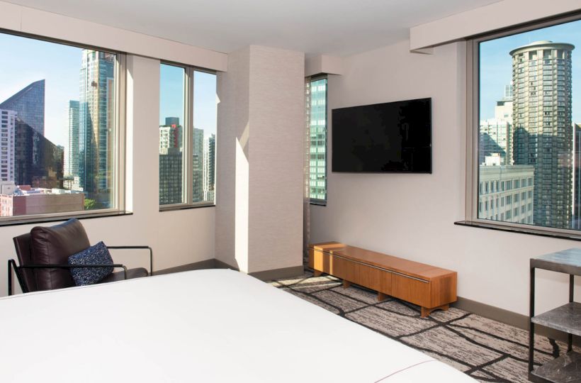 A modern hotel room with large windows shows a city skyline. There's a bed, TV, chair, and a small table in the room.
