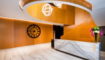 A modern lobby with a marble reception desk, large wall clock, spiral wooden paneling, and hanging lights, adorned with a flower arrangement.