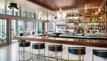 It's a modern bar with a sleek wooden counter, stylish stools, and shelves full of bottles, situated in a bright, open space.