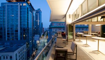 A modern, outdoor rooftop terrace with seating overlooks a city street lined with tall buildings at dusk.