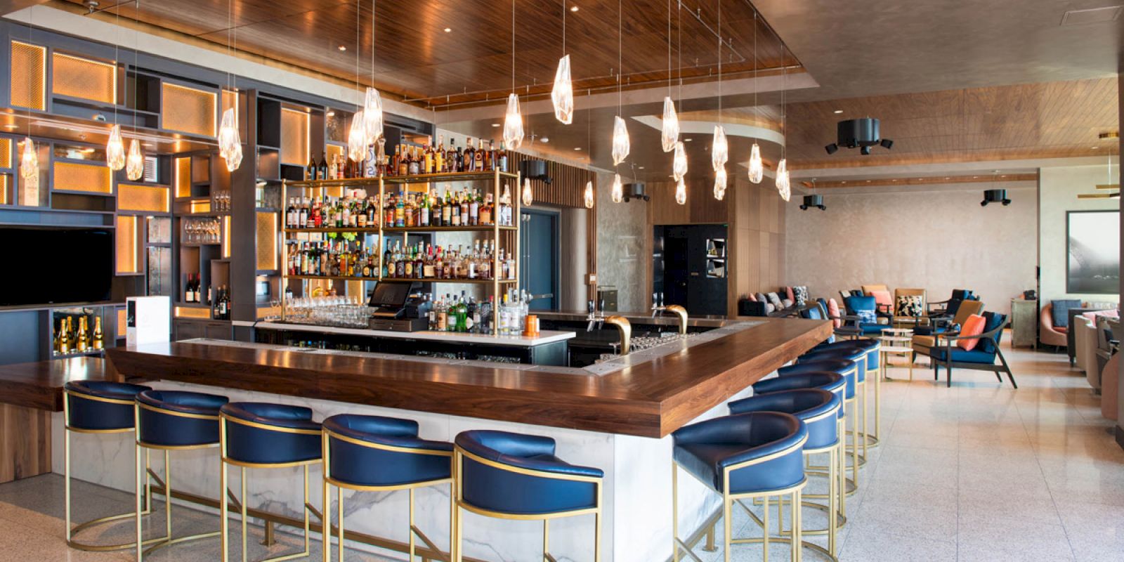 A modern bar with blue stools, a wooden counter, and a well-stocked shelf of bottles. Hanging lights and a cozy seating area complete the scene.