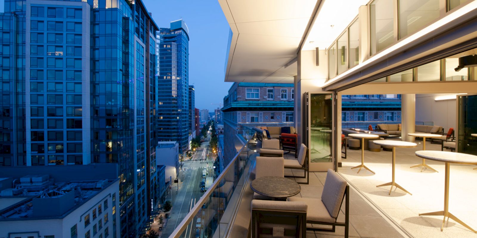 A modern balcony scene at dusk overlooking a city street with tall buildings and tables and chairs on the patio.