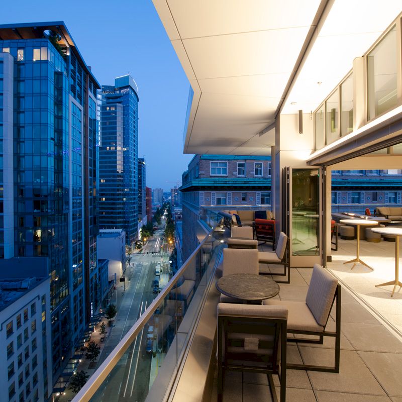 A modern balcony at dusk overlooks a city street flanked by tall buildings, with seating areas and ambient lighting visible.