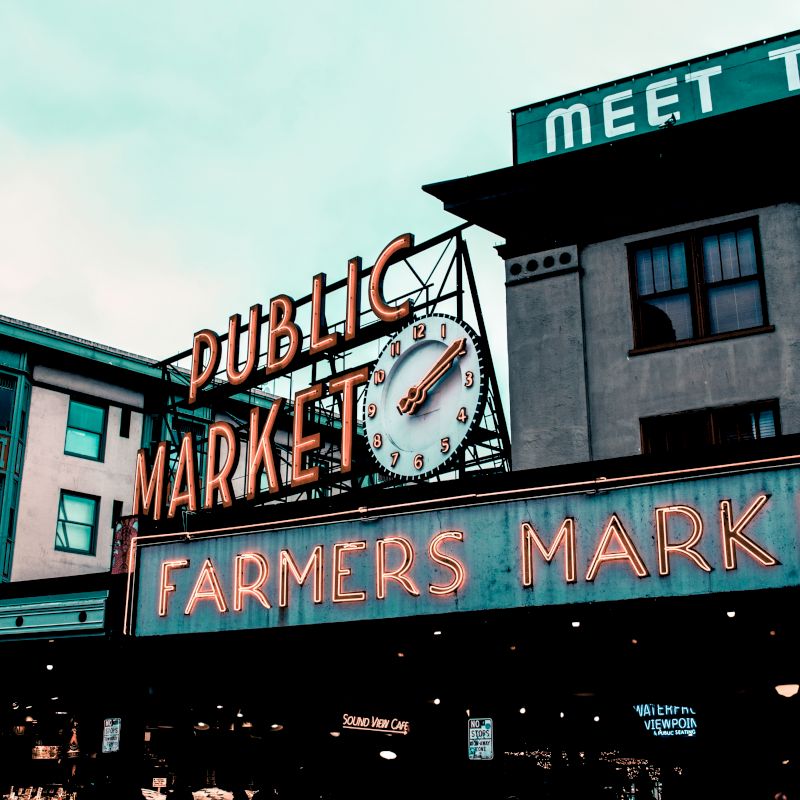 The image shows a famous farmers market with vintage signs and a clock on the building facade. The signs read "PUBLIC MARKET" and "FARMERS MARKET."