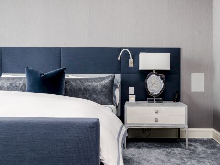 A modern bedroom with a navy bed, white bedding, and a bedside table. The table has a lamp and geode decor, with a cozy carpeted floor.
